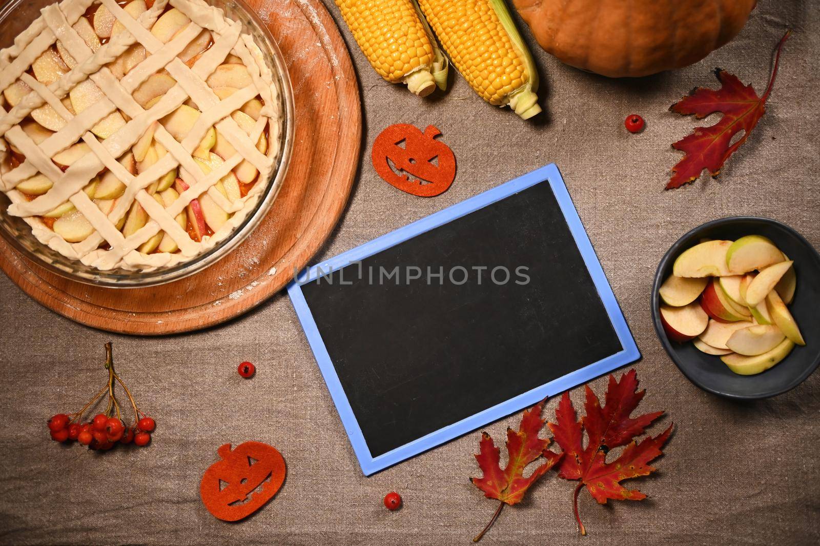 Flat lay. A blackboard with copy ad space, Thanksgiving apple pie with crispy crust lattice, harvested crop of pumpkin and corn next to autumn maple leaves and viburnum berries on a linen tablecloth