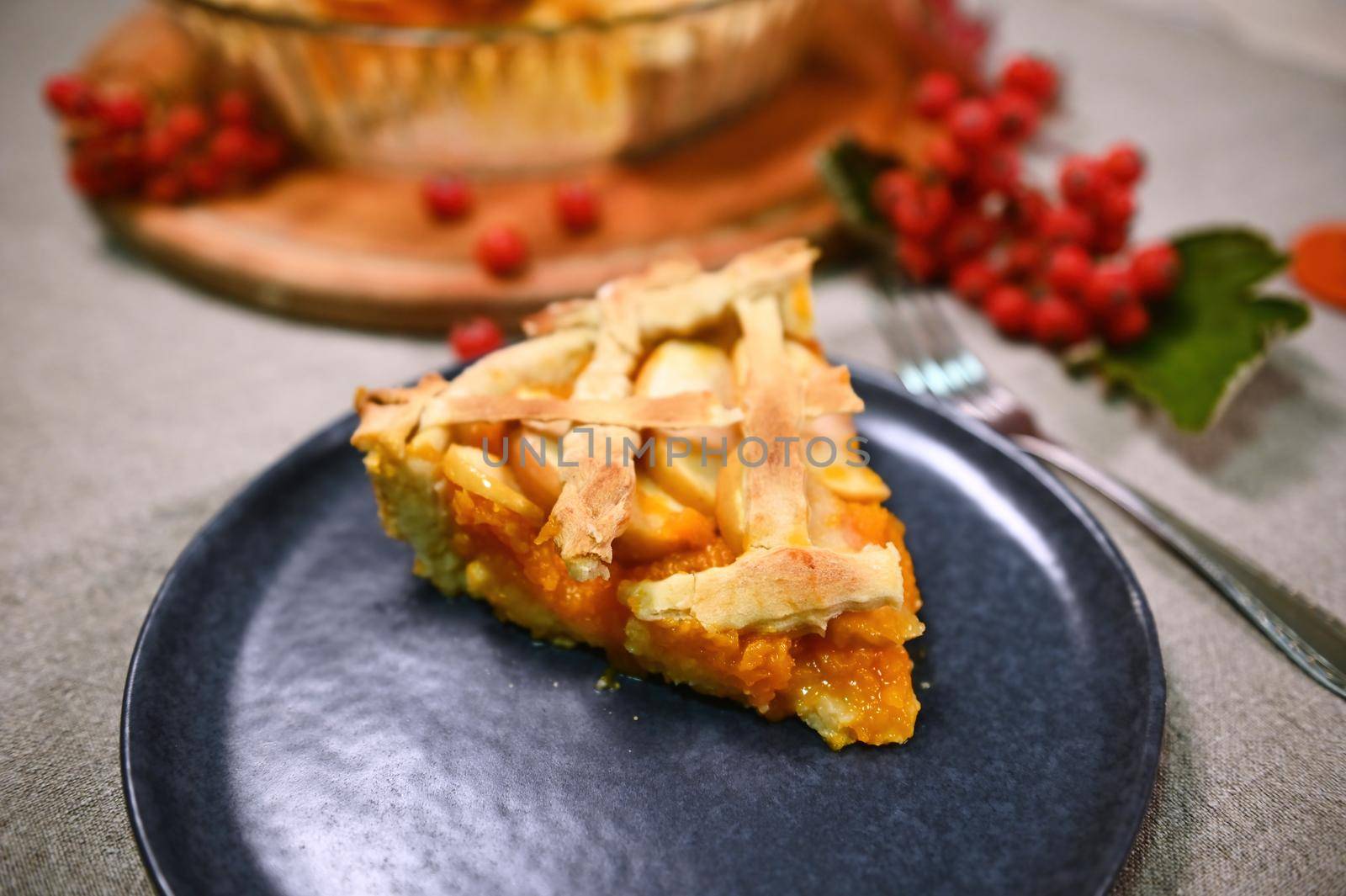 Sweet homemade pumpkin pie with caramelized apples and crispy crust lattice ona blue ceramic plate, with blurred baking dish with Thanksgiving cake and red autumnal viburnum berries on the background