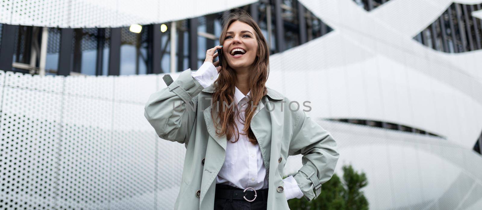 a young woman speaks on a mobile phone with a wide smile on her face against the background of an aluminum facade of an office building by TRMK