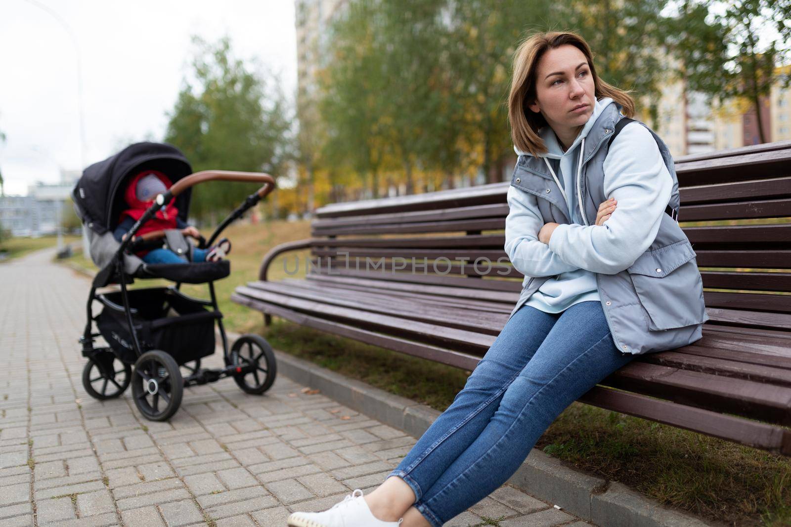 upset mother sits on a bench separated from the pram with her baby, the concept of postpartum depression by TRMK