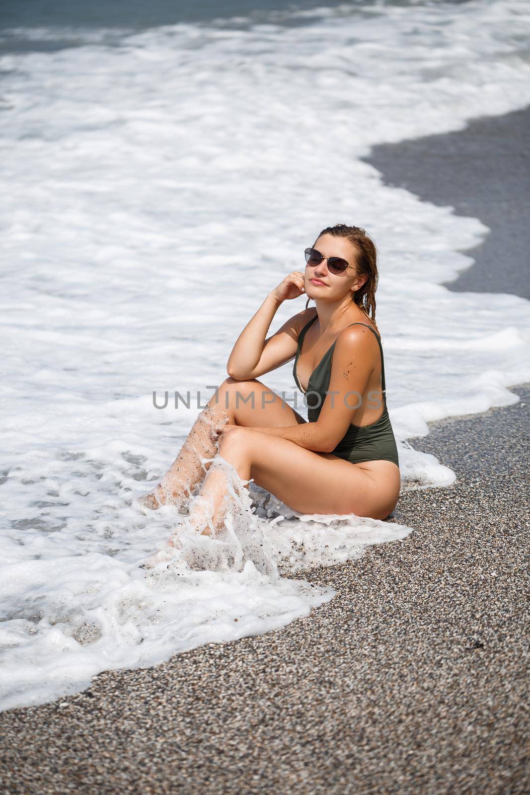 Woman in sunglasses in a green swimsuit stands on the sand by the ocean and has a rest