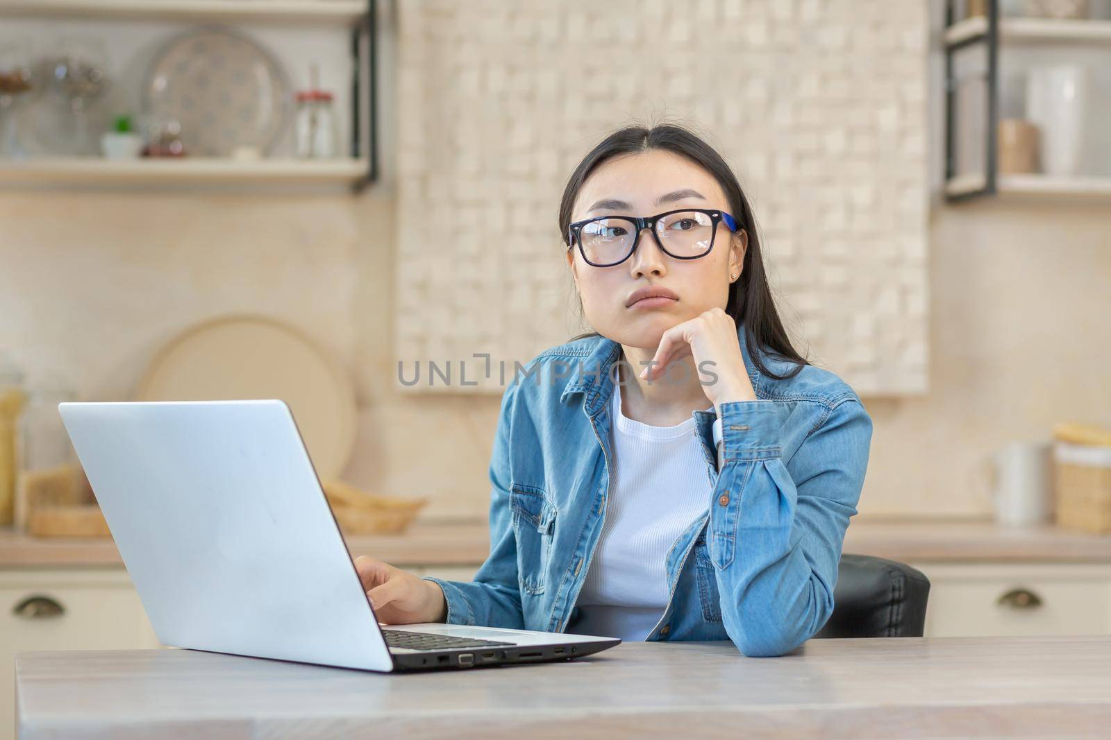 Sad and upset woman sitting at home in the kitchen, asian woman with laptop working online remotely, depressed student,