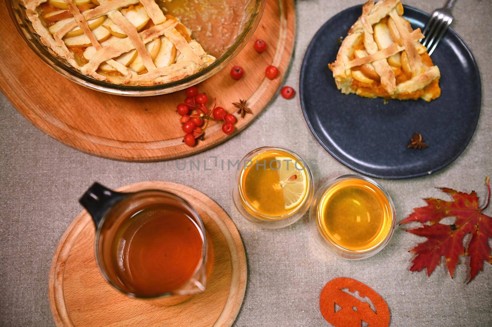 Tea time. Still life with freshly baked homemade pumpkin pie with crispy crust for Thanksgiving, teapot and two cups of hot drink surrounded by fallen autumn maple leaves on a linen tablecloth