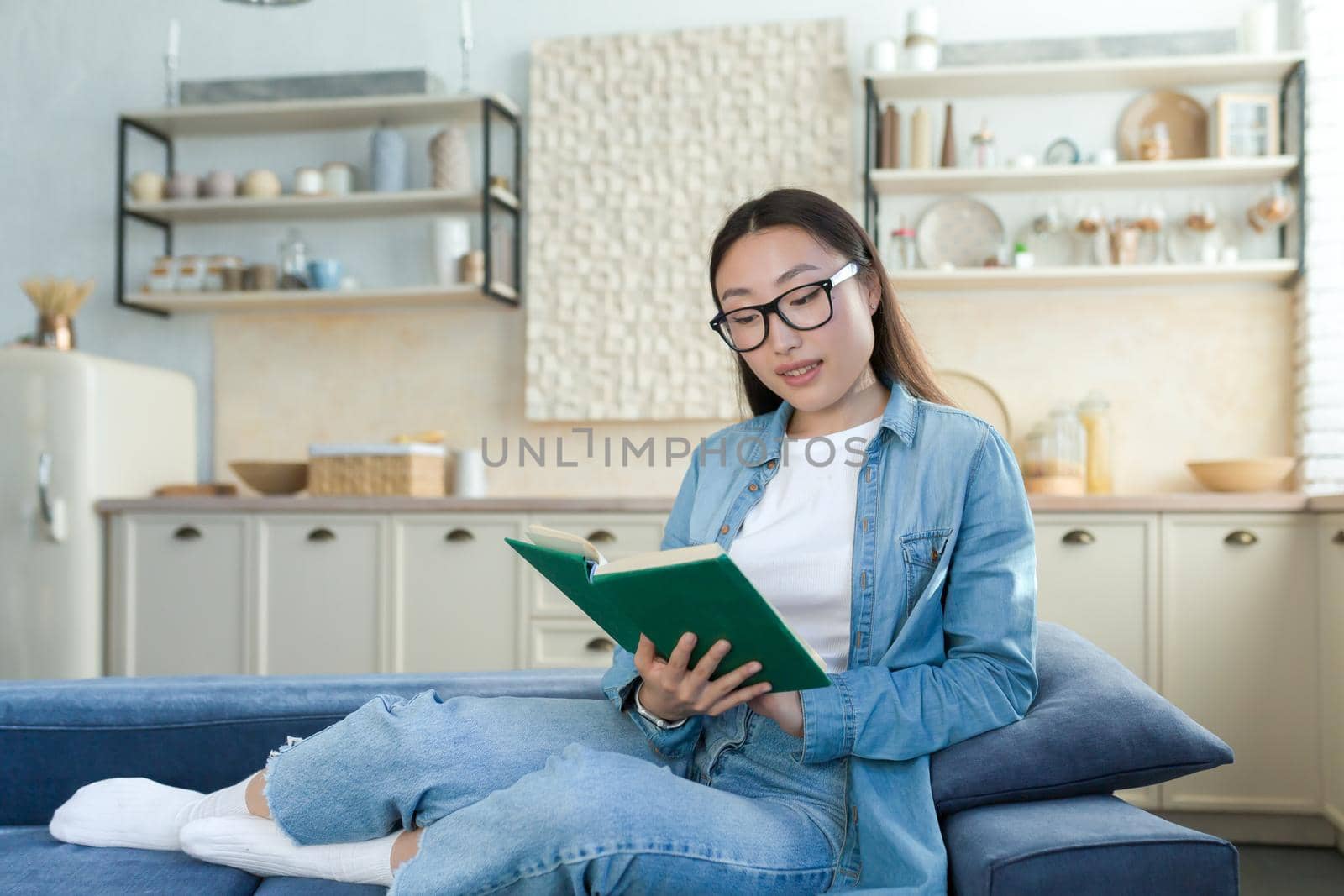 Young beautiful asian woman in glasses and denim shirt studying at home, happy female student smiling reading book sitting on sofa in living room.