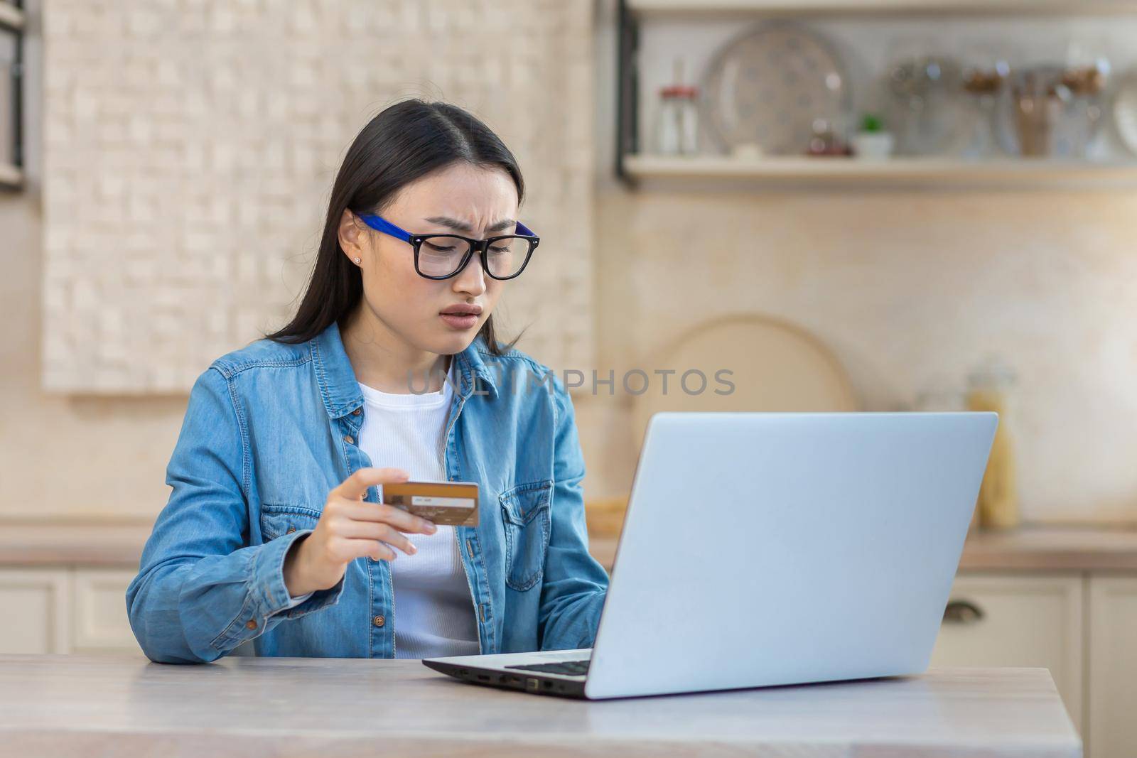 Upset and sad woman in depression trying to make online purchase in online store, Asian woman sitting at home in kitchen using laptop and bank credit card, unable to make payment online by voronaman