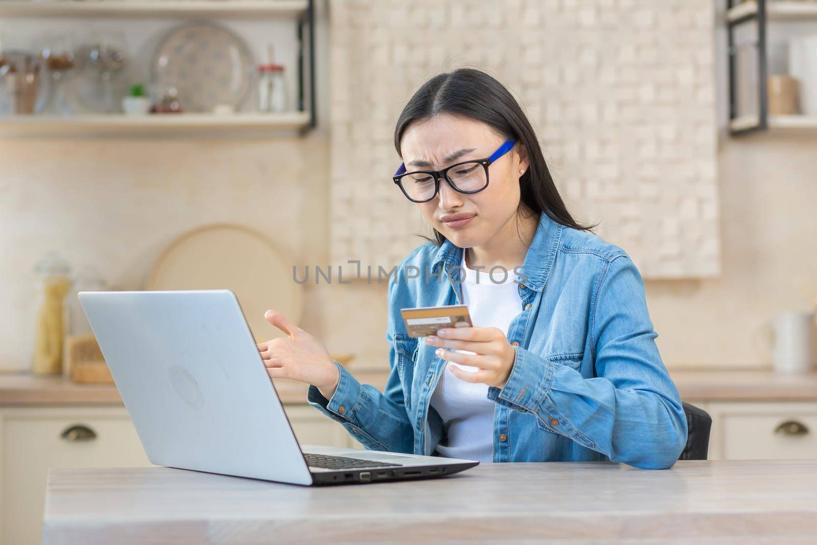 Upset and sad woman in depression trying to make online purchase in online store, Asian woman sitting at home in kitchen using laptop and bank credit card, unable to make payment online by voronaman