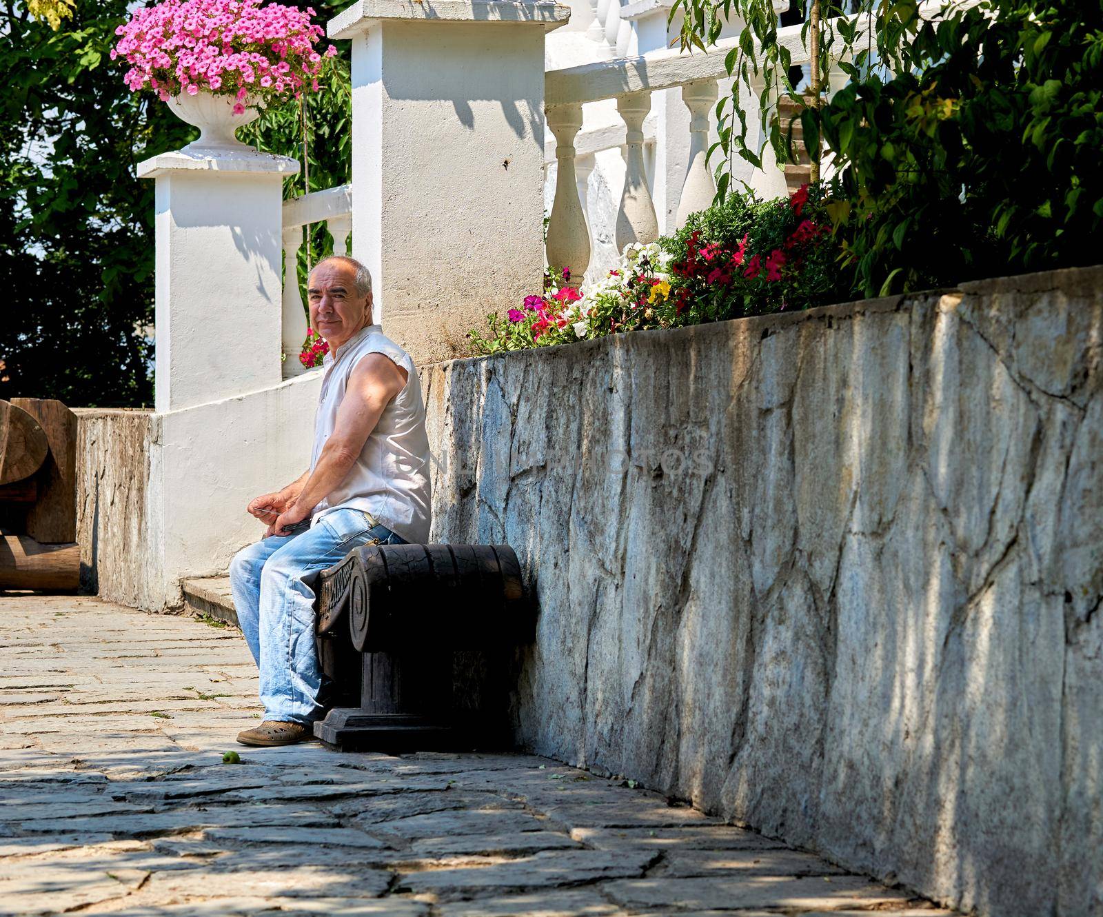 Elderly man resting on a bench near a white vintage stone wall in a city park by jovani68