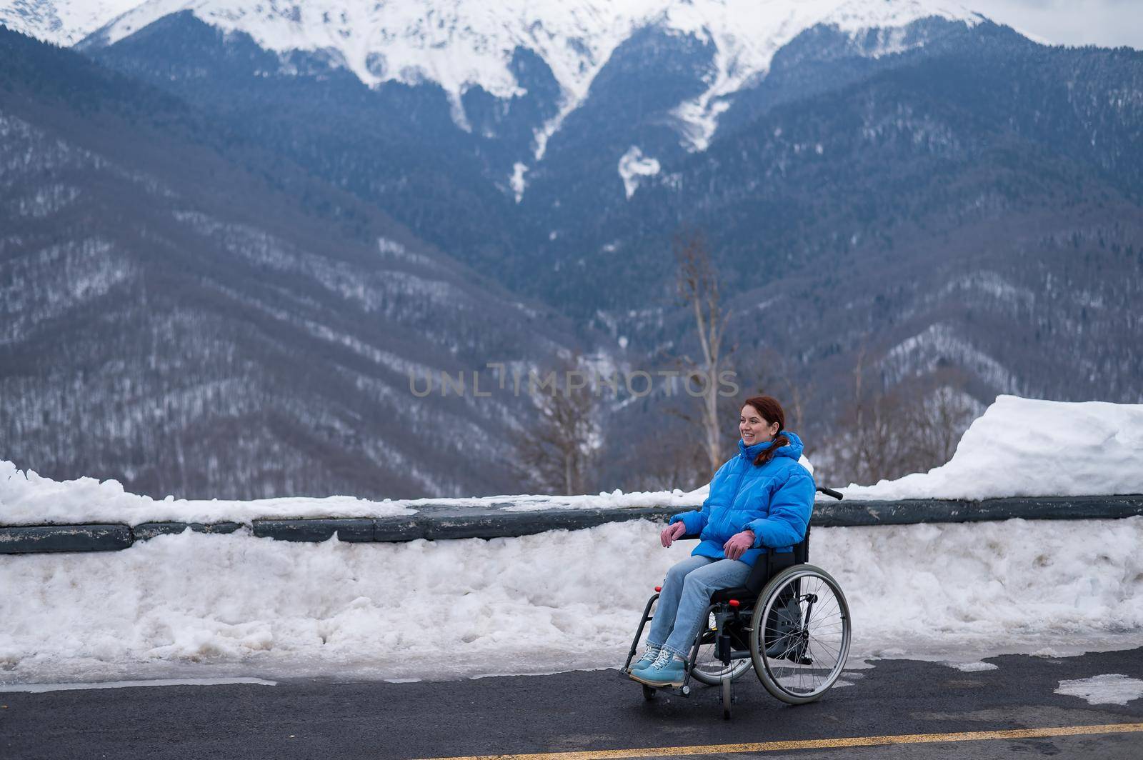 Smiling woman dressed in a blue coat sits in a wheelchair on a point view looks at high mountains