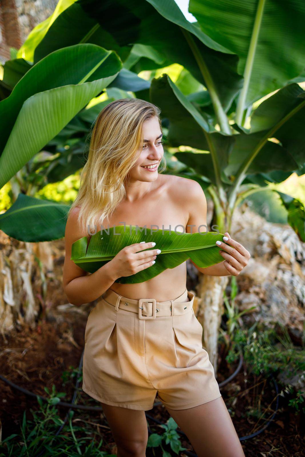 A beautiful young woman stands in a beautiful pose near a banana tree, her little girl covers her breasts with a leaf on a banana plantation. by Dmitrytph