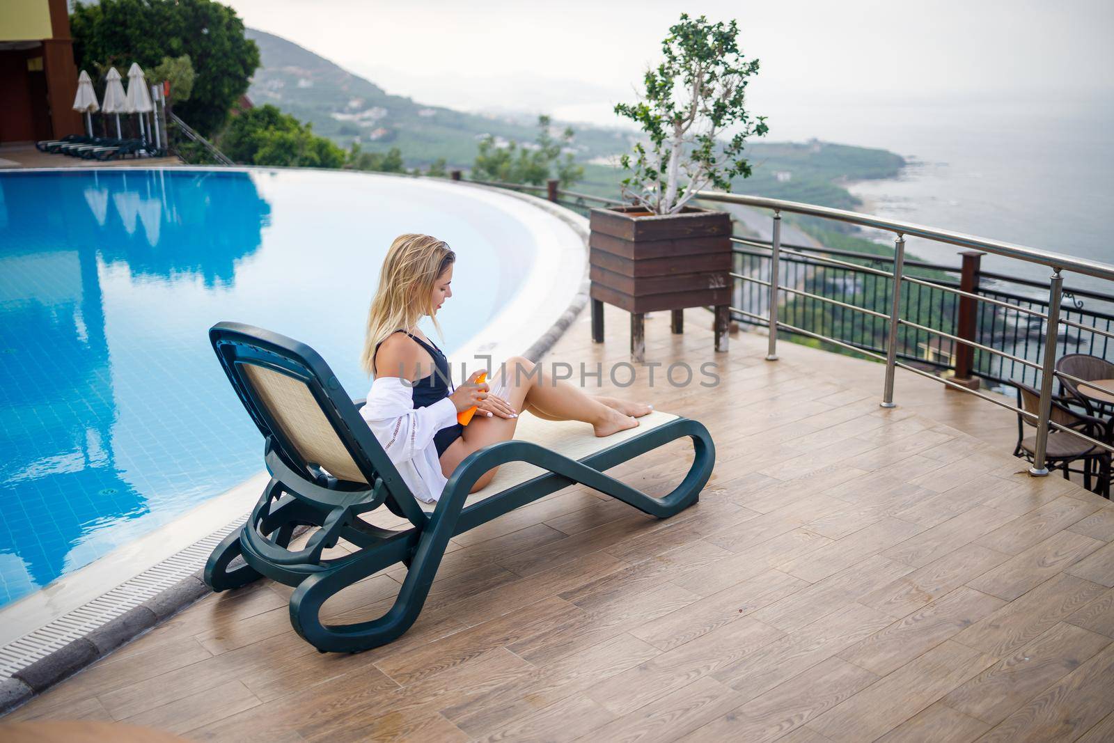 A beautiful young woman in a swimsuit and a white shirt sits on a sun lounger by the pool and rubs her body with sunscreen. Summer skin care, protection against skin burning