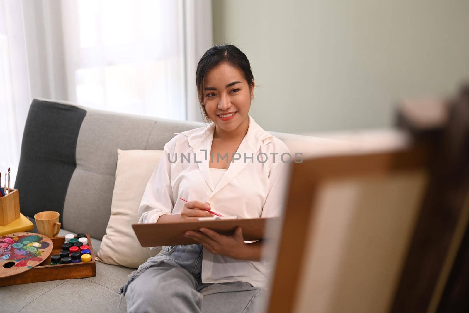Pleasant millennial woman sitting on couch in bright living room and painting on canvas. Art and leisure activity concept by prathanchorruangsak