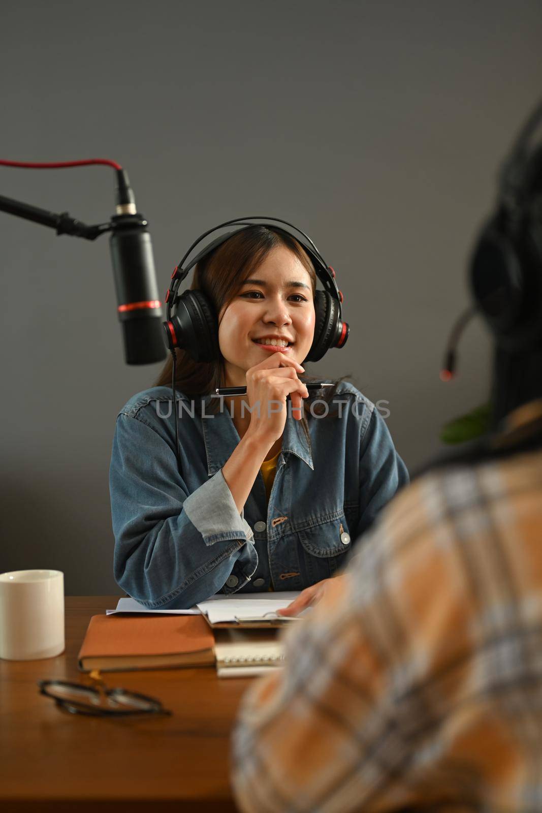 Friendly radio host in headphone discussing various topics with her guest while streaming live audio podcast from home studio by prathanchorruangsak