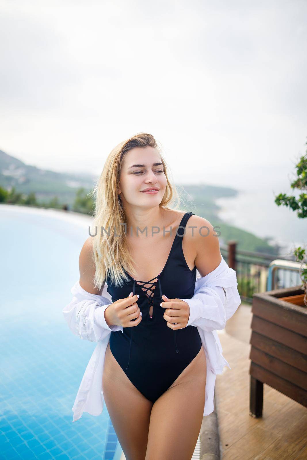 Beautiful woman in black swimsuit posing near outdoor pool with sea view