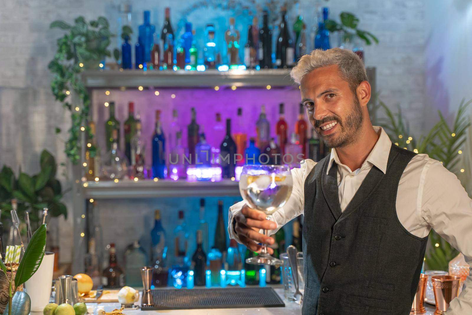 Elegant barman smiling on bar counter with a cocktail in his hand. Toasting in the air. by PaulCarr