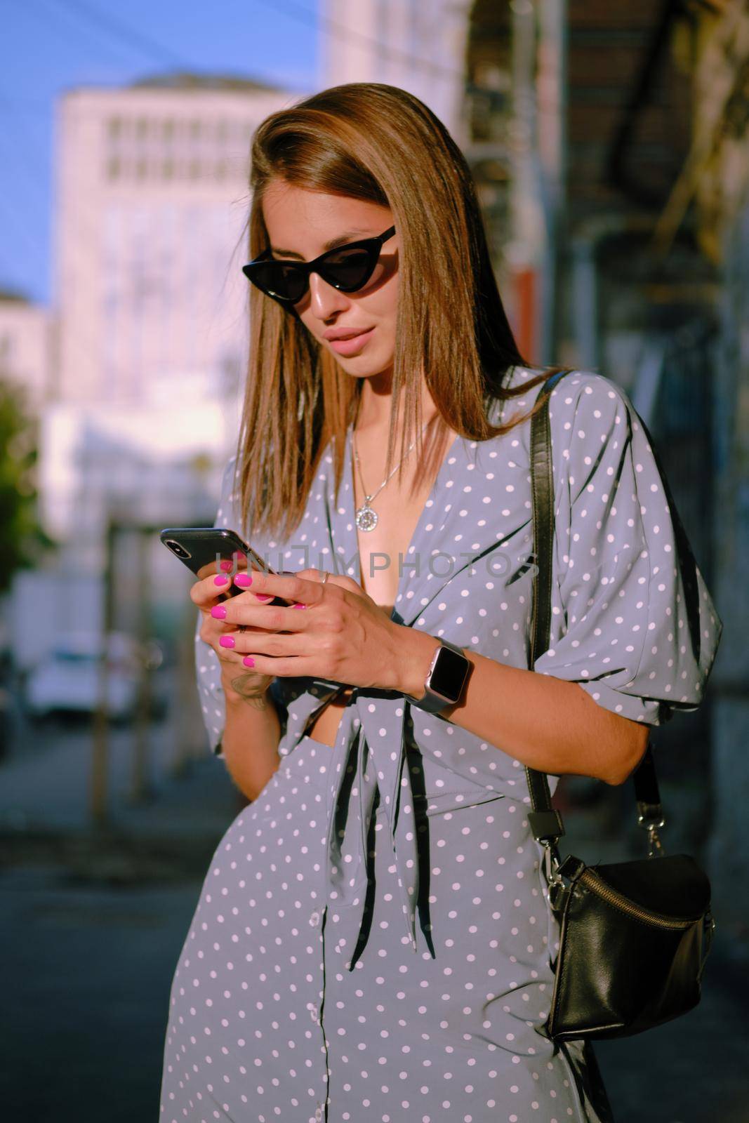 Graceful blonde maiden in a long blue dress with polka-dots, watch, sunglasses, with a pendant around her neck and a small black handbag on her shoulder is using her smartphone while walking alone in the city. The concept of fashion and style. Close-up shot.