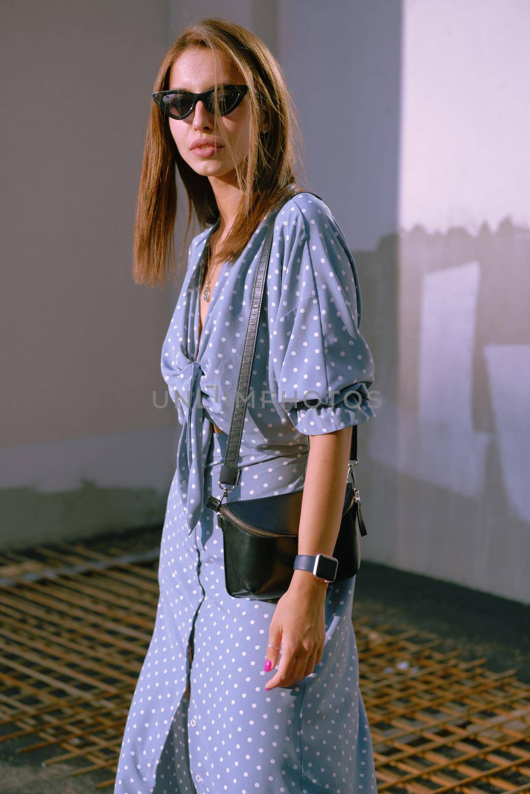 Beautiful blonde female in a long blue dress with polka-dots, watch, sunglasses, with a pendant around her neck and a small black handbag on her shoulder is looking at the camera while walking alone in the city. The concept of fashion and style. Close-up shot.