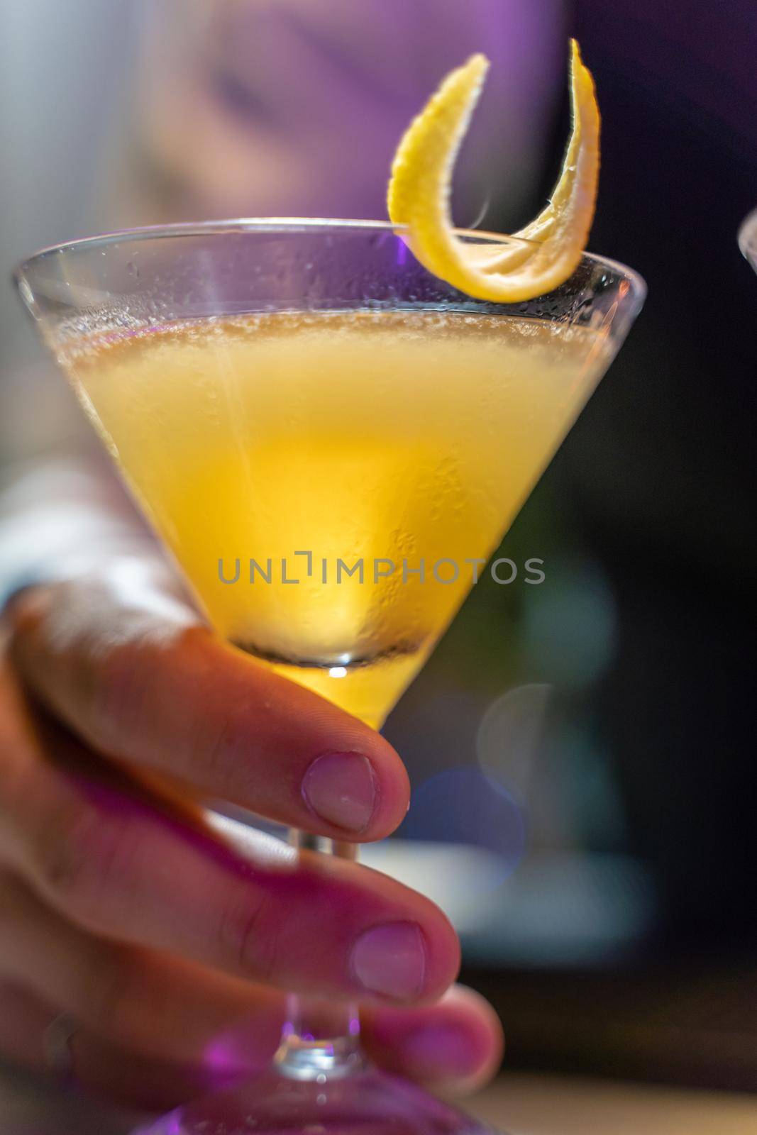 Close-up of a barman's hand serving a beautiful yellow cocktail with lemon peel on the bar counter in the nightclub. by PaulCarr