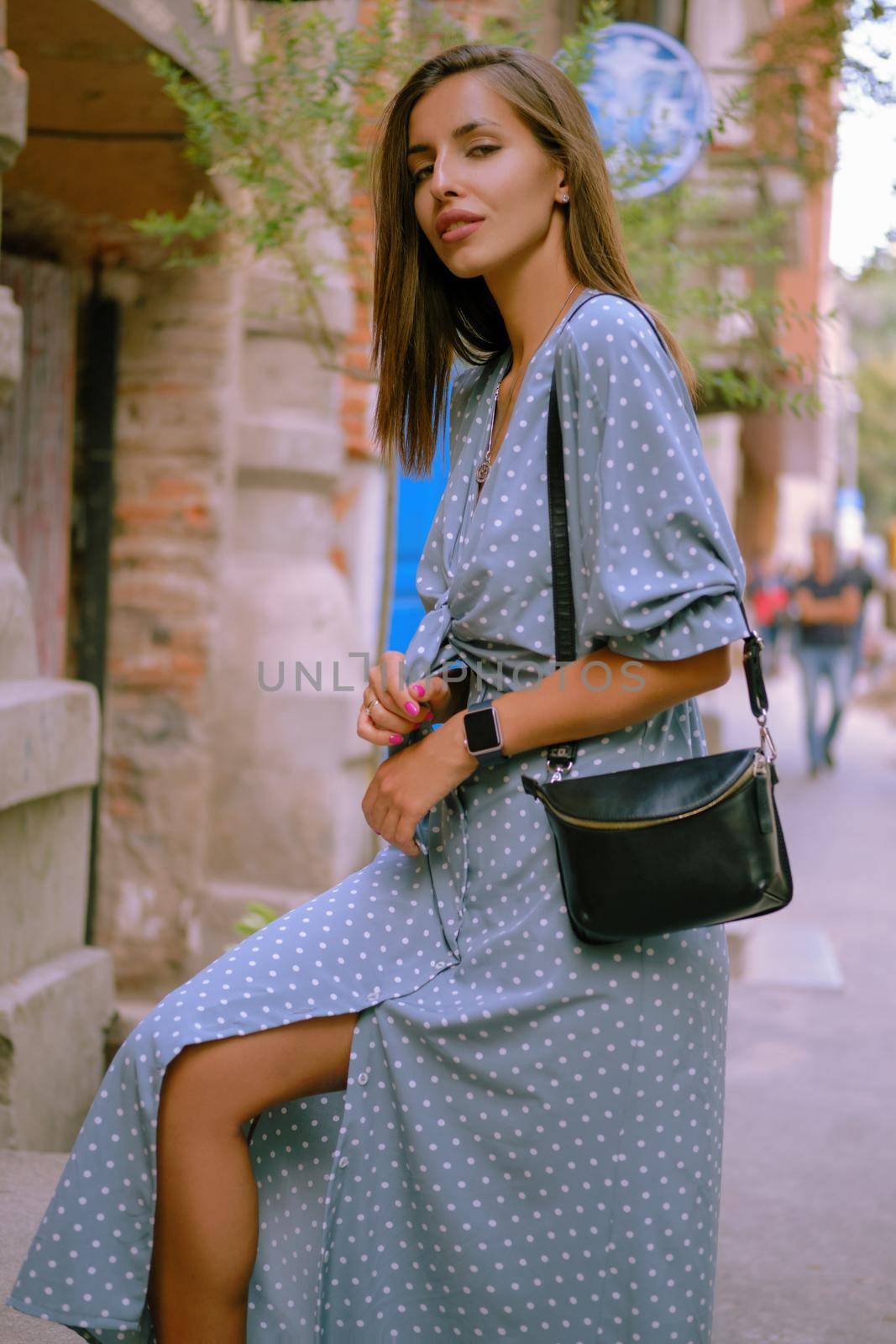 Soft Focus. Alluring blonde lady in a long blue dress with polka-dots, watch, with a pendant around her neck and a small black handbag on her shoulder is looking at the camera while posing standing sideways, walking alone in the city. The concept of fashion and style. Close-up shot.