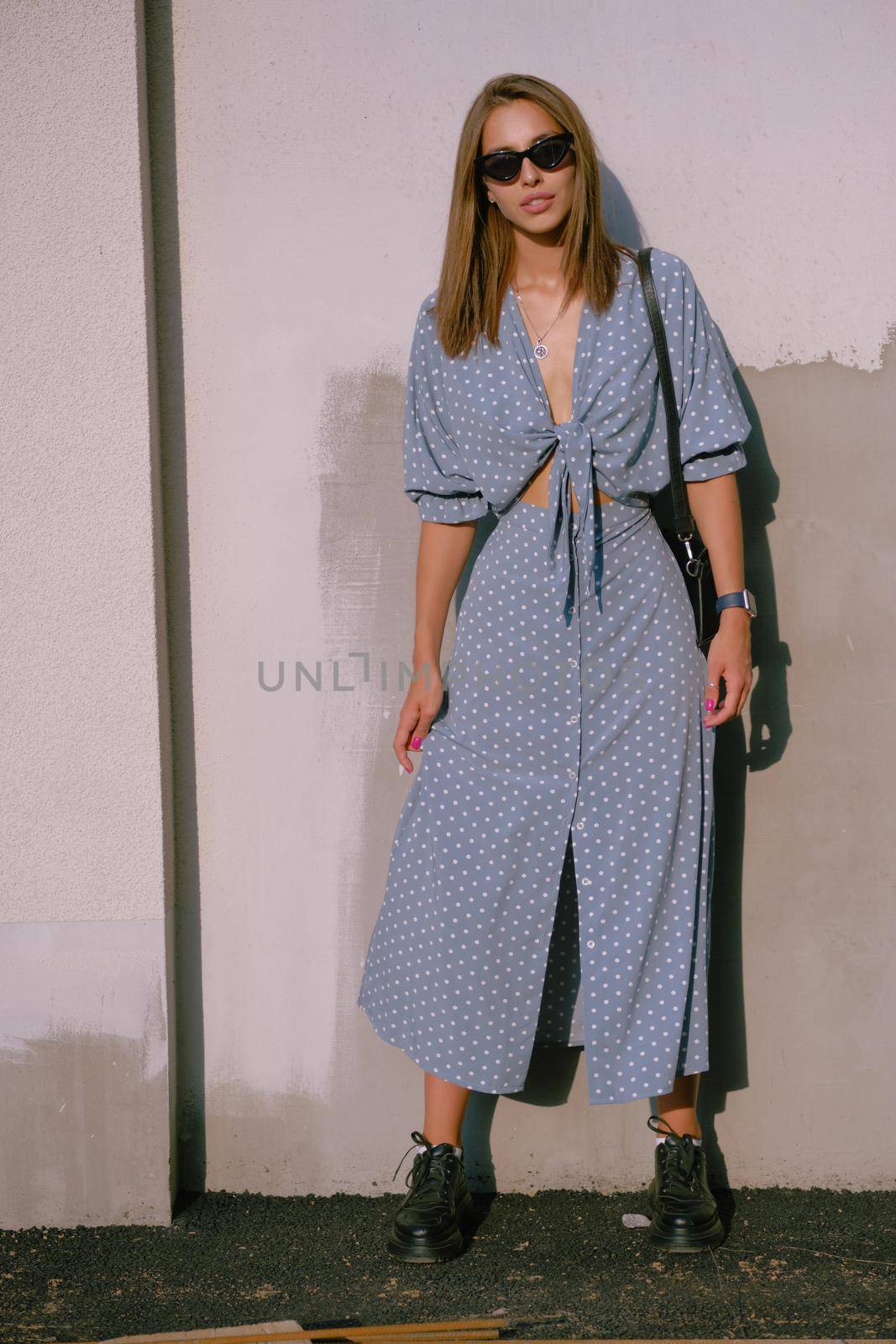 Gorgeous blonde woman in a long blue dress with polka-dots, black boots, watch, sunglasses, with a pendant around her neck and a small handbag on her shoulder is posing near a white wall while walking alone in the city. The concept of fashion and style. Full length shot.