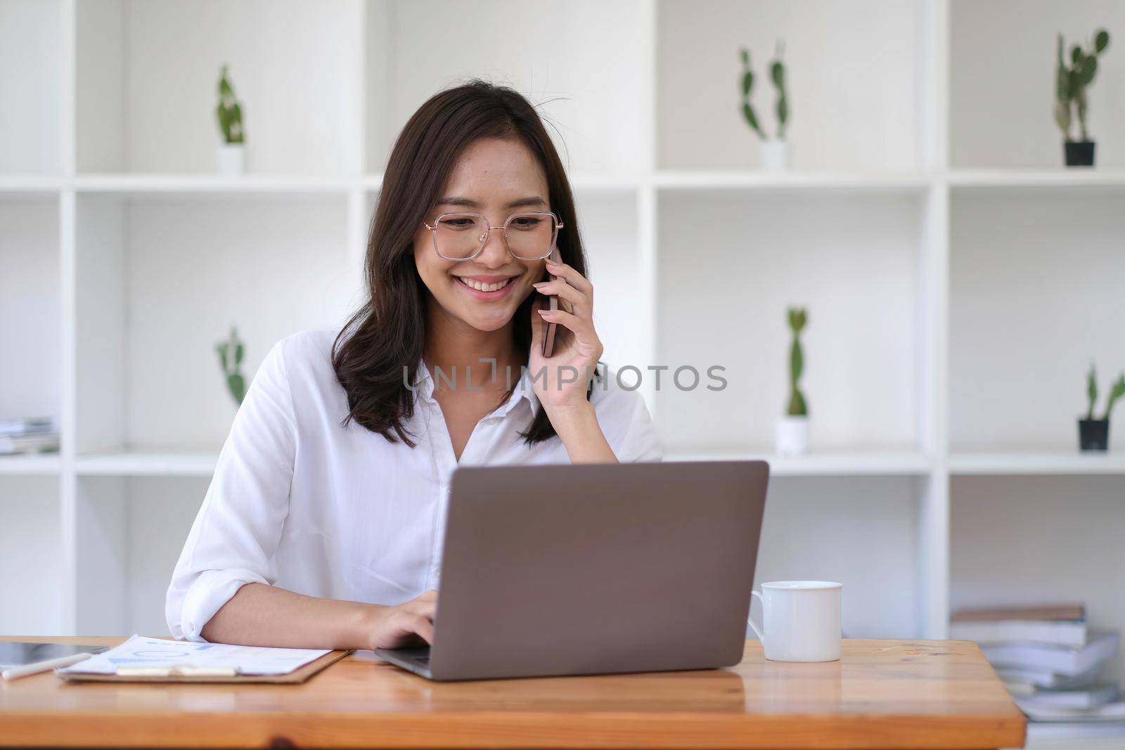 Smiling beautiful young Asian business woman enjoying phone call with customer and commenting at the office while working on a laptop computer. by wichayada