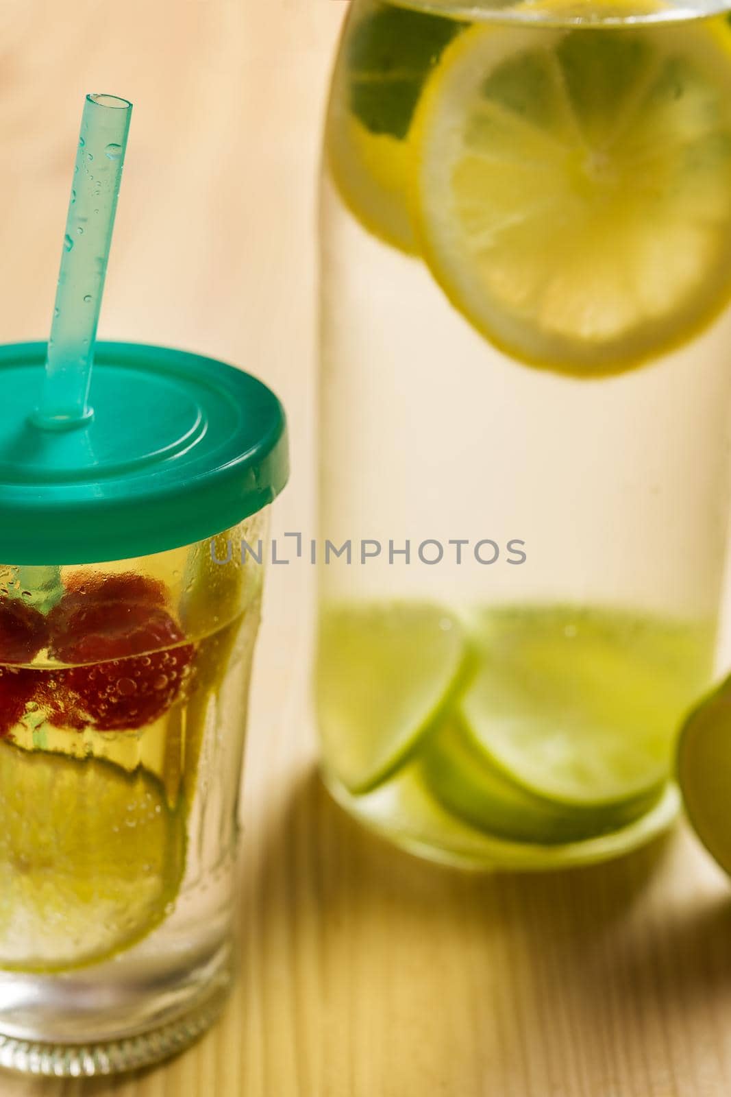 vertical photo of bottle and glass with cold water and slices of lime, lemon and red berries, illuminated by sunlight on a wooden table with some pieces of citrus, summer refreshments background