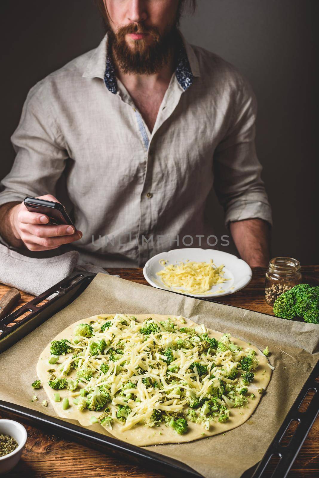 Bearded man surfing phone while cooking pizza with broccoli, pesto sauce, spices and cheese