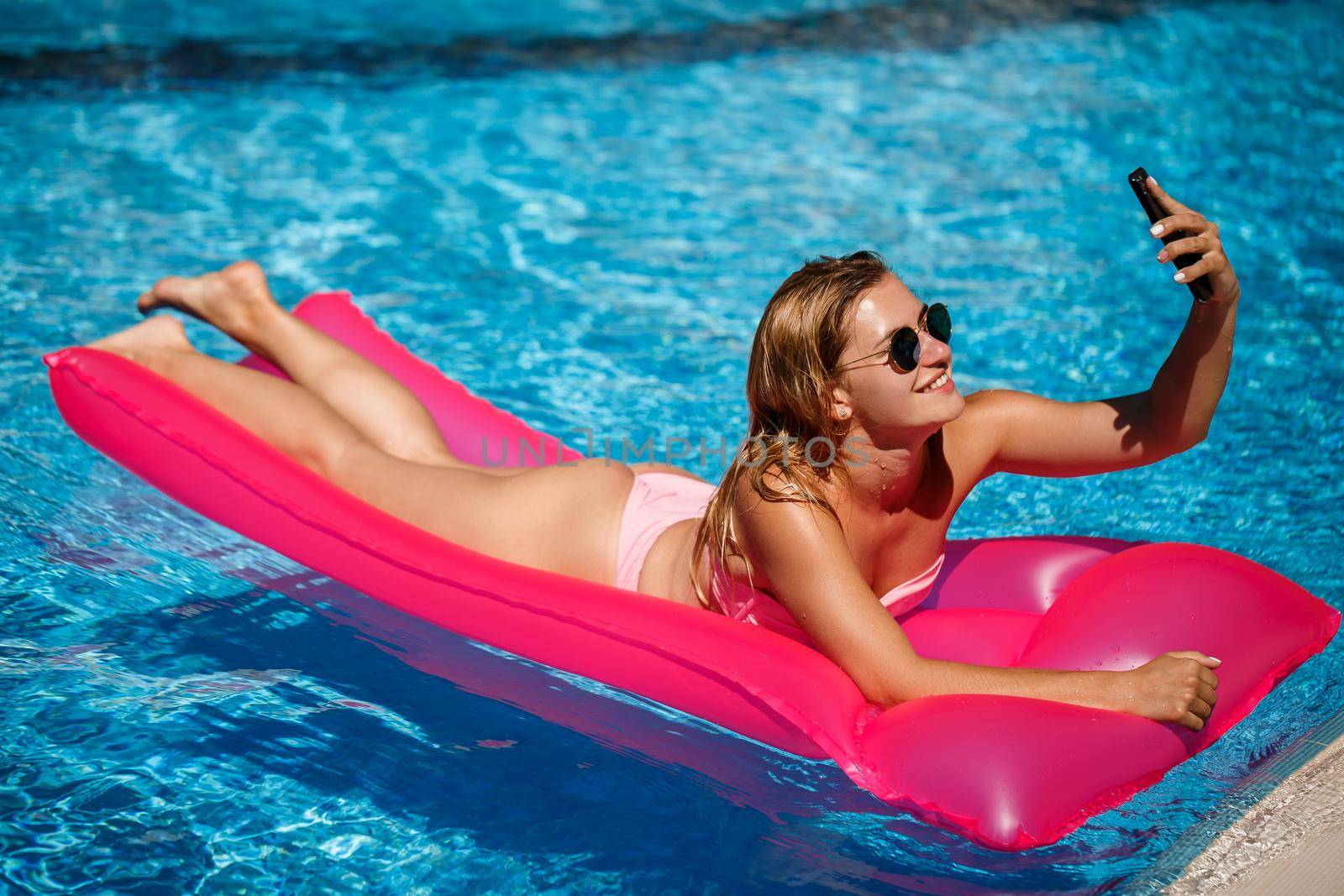 Sexy woman with a phone in a swimsuit lies on a pink inflatable mattress in the pool. Relax by the pool on a hot summer sunny day. Vacation concept by Dmitrytph