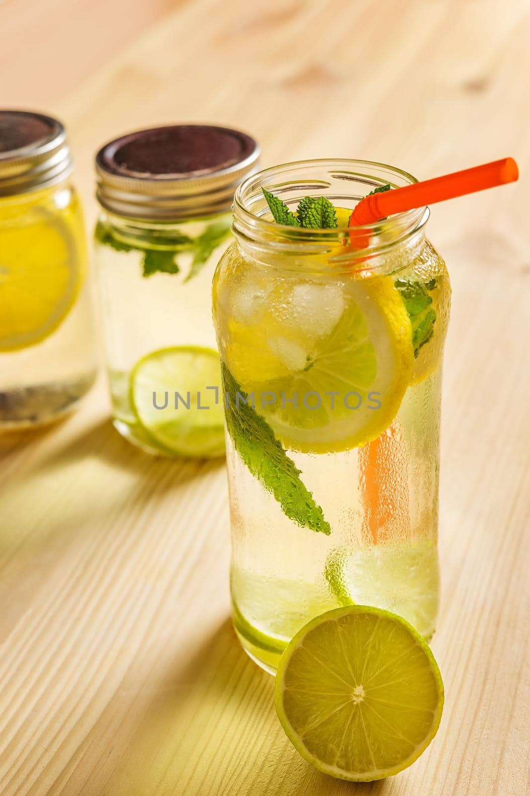 vertical image os a glass jar full of water with ice, slices of lemon, lime and mint leaves, also has a cane to drink and another lemonade unfocused in the background, all is on a wooden table