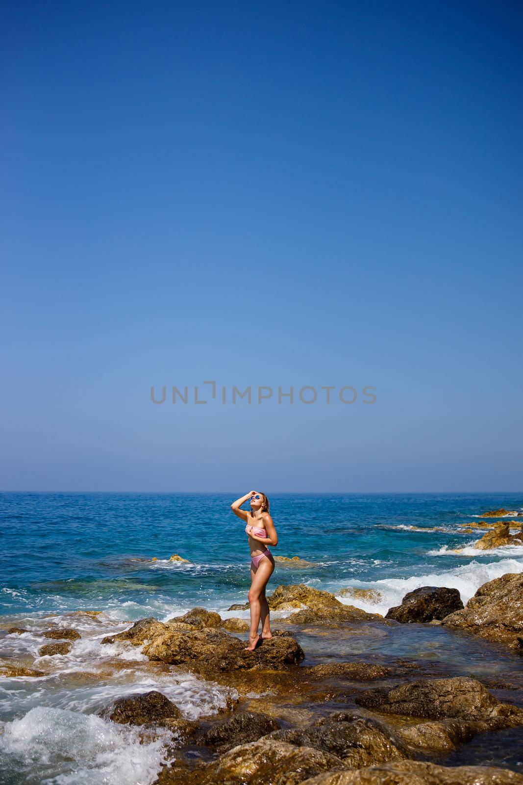 Beautiful young woman in a swimsuit on a rocky beach on a sunny day against the backdrop of waves. Vacation in the summer season. Selective focus by Dmitrytph