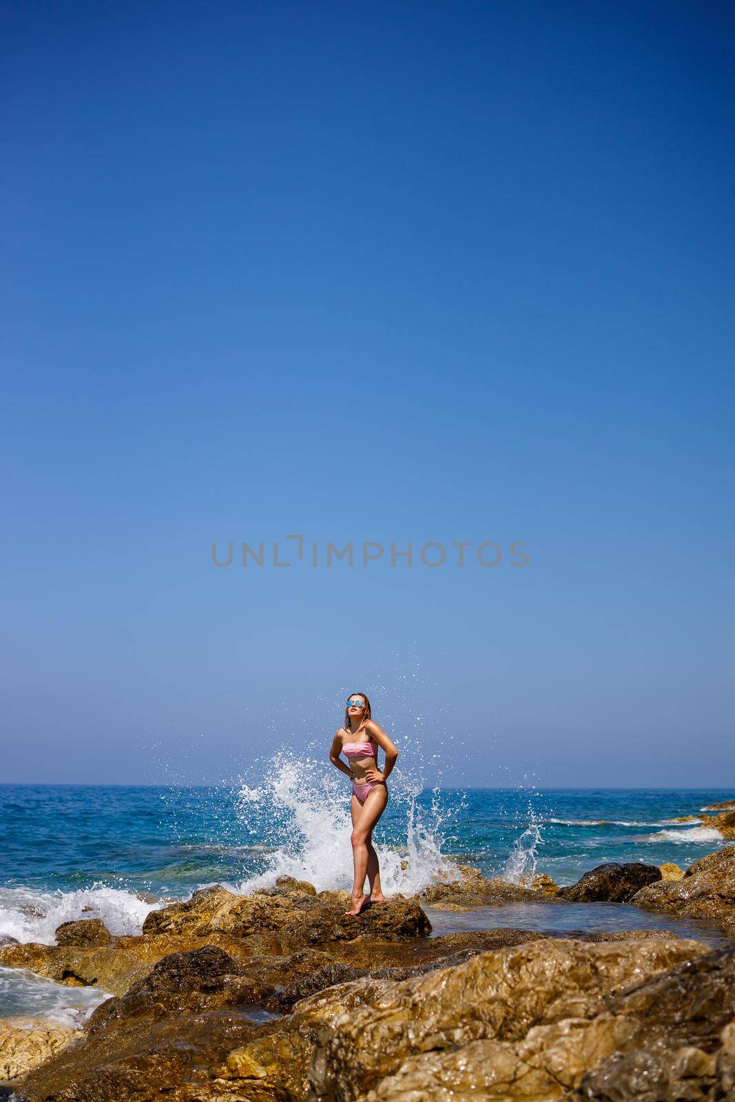 Beautiful young woman in a swimsuit on a rocky beach on a sunny day against the backdrop of waves. Vacation in the summer season. Selective focus by Dmitrytph