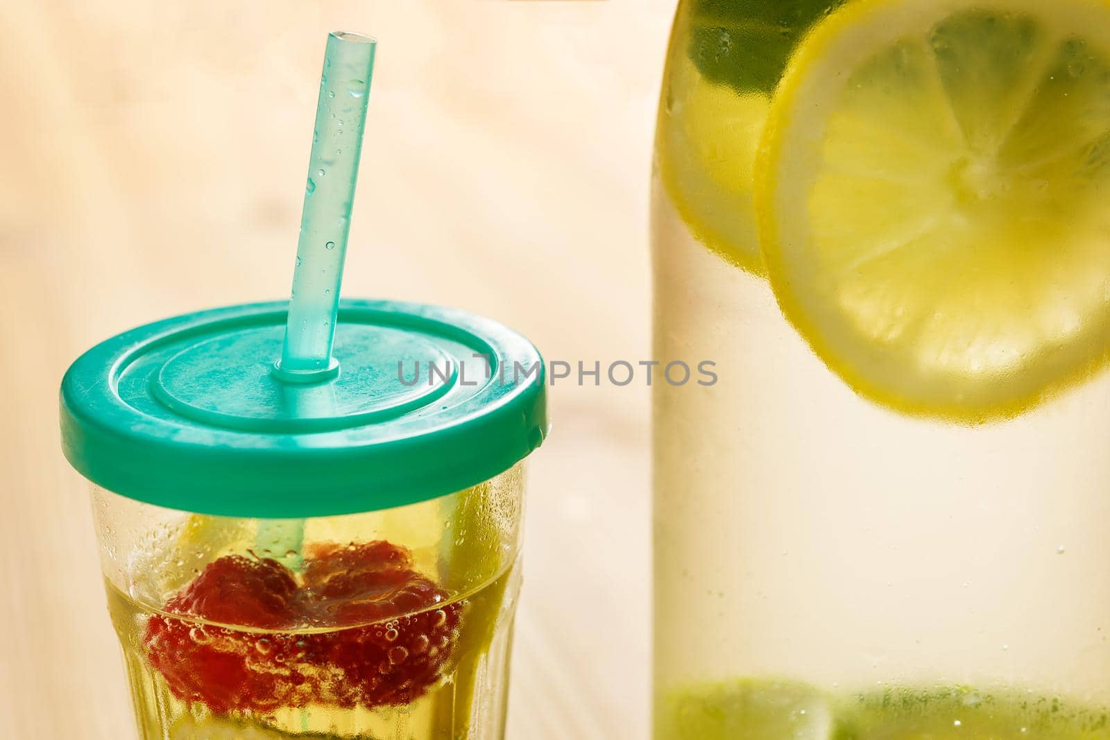 glass with lid and cane and a bottle, with cold water and slices of lemon, lime, berries and mint, are illuminated by sunlight on a wooden table with some pieces of citrus