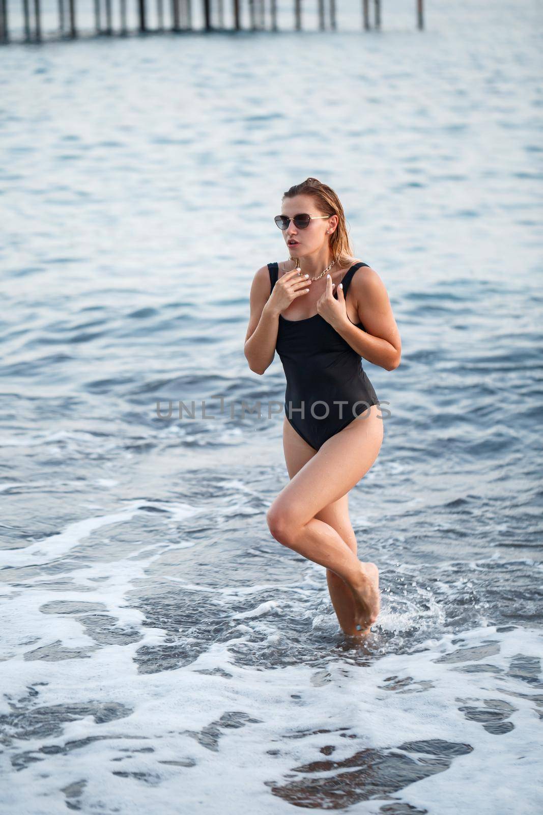 Portrait of a beautiful woman in a black swimsuit with blond hair posing on the beach by the sea. Young woman walking on the beach