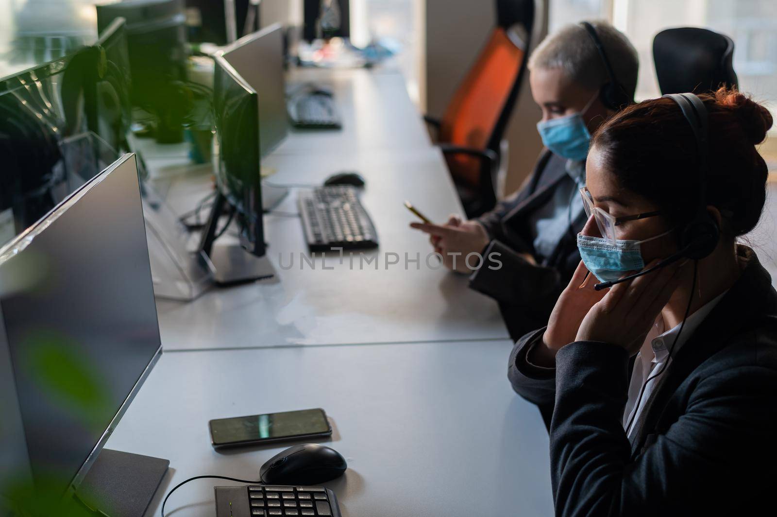 Two women in medical masks and headsets are working in the office.