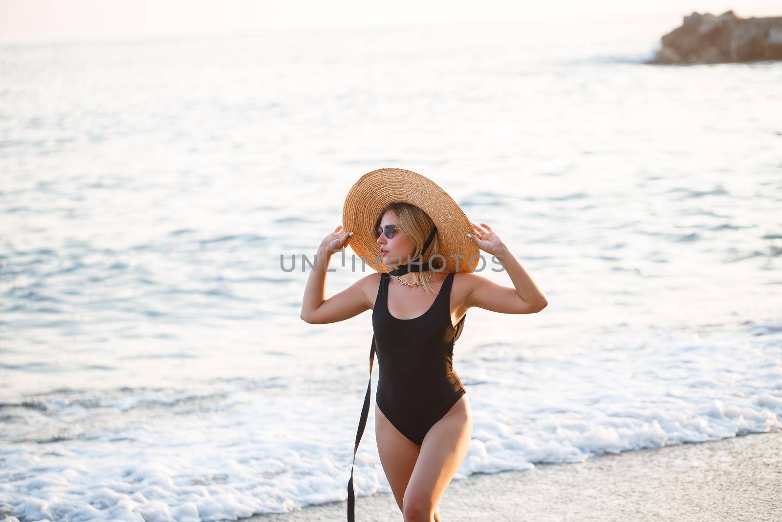 Beautiful girl in a black swimsuit and hat on a sandy beach at the sea in the sunset sunlight