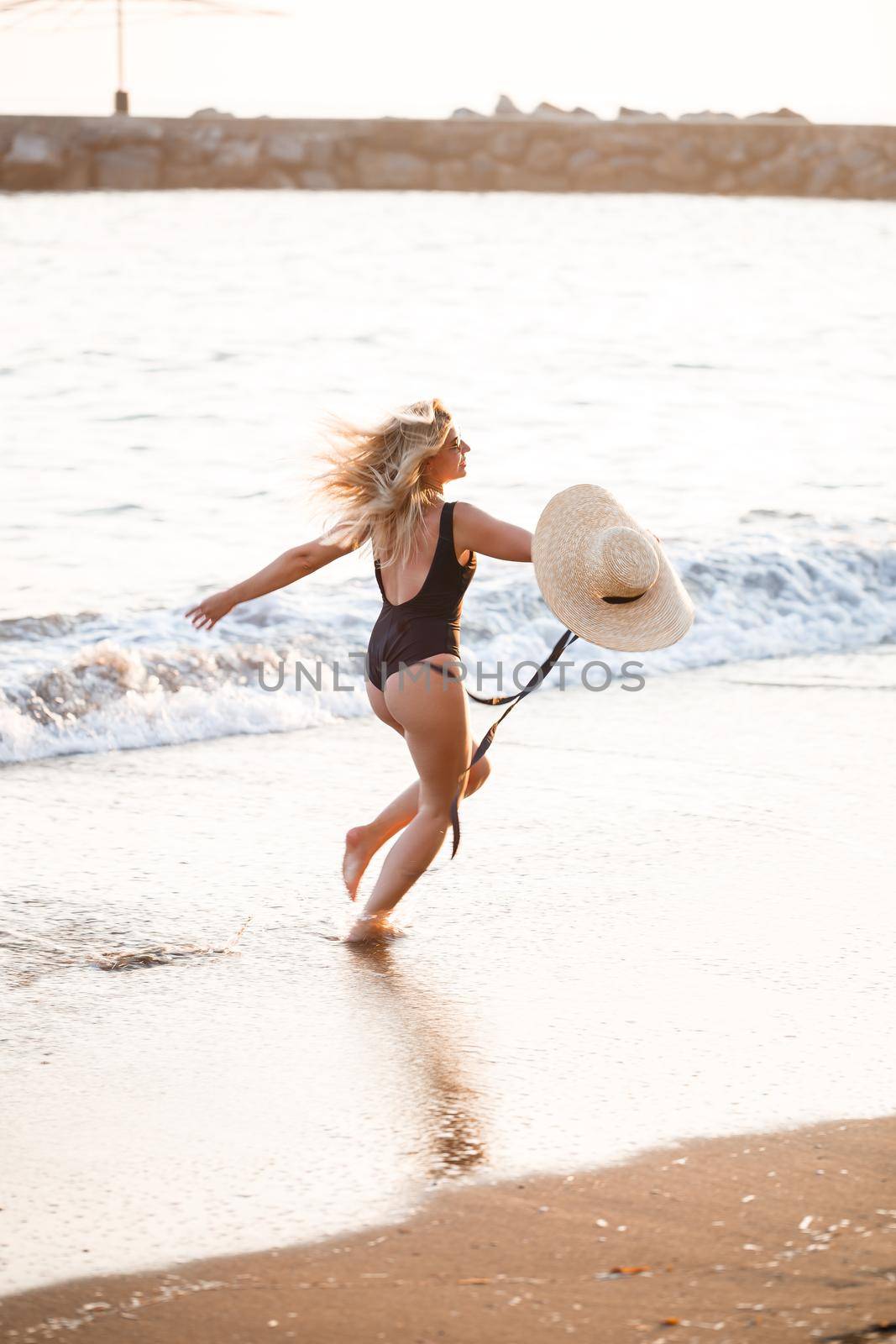 Beautiful girl in a black swimsuit and hat on a sandy beach at the sea in the sunset sunlight