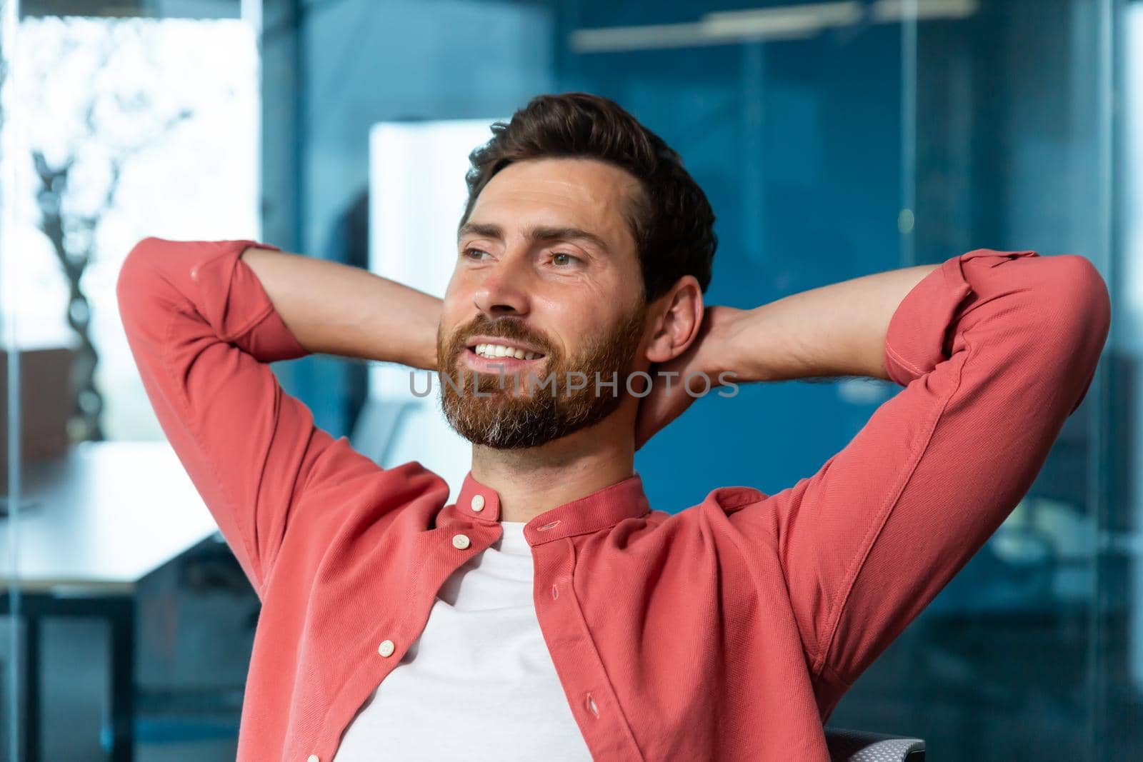 Young successful businessman startuper rests in office, man in red shirt throws hands behind head and dreams visualizes, designer thinks about new project.