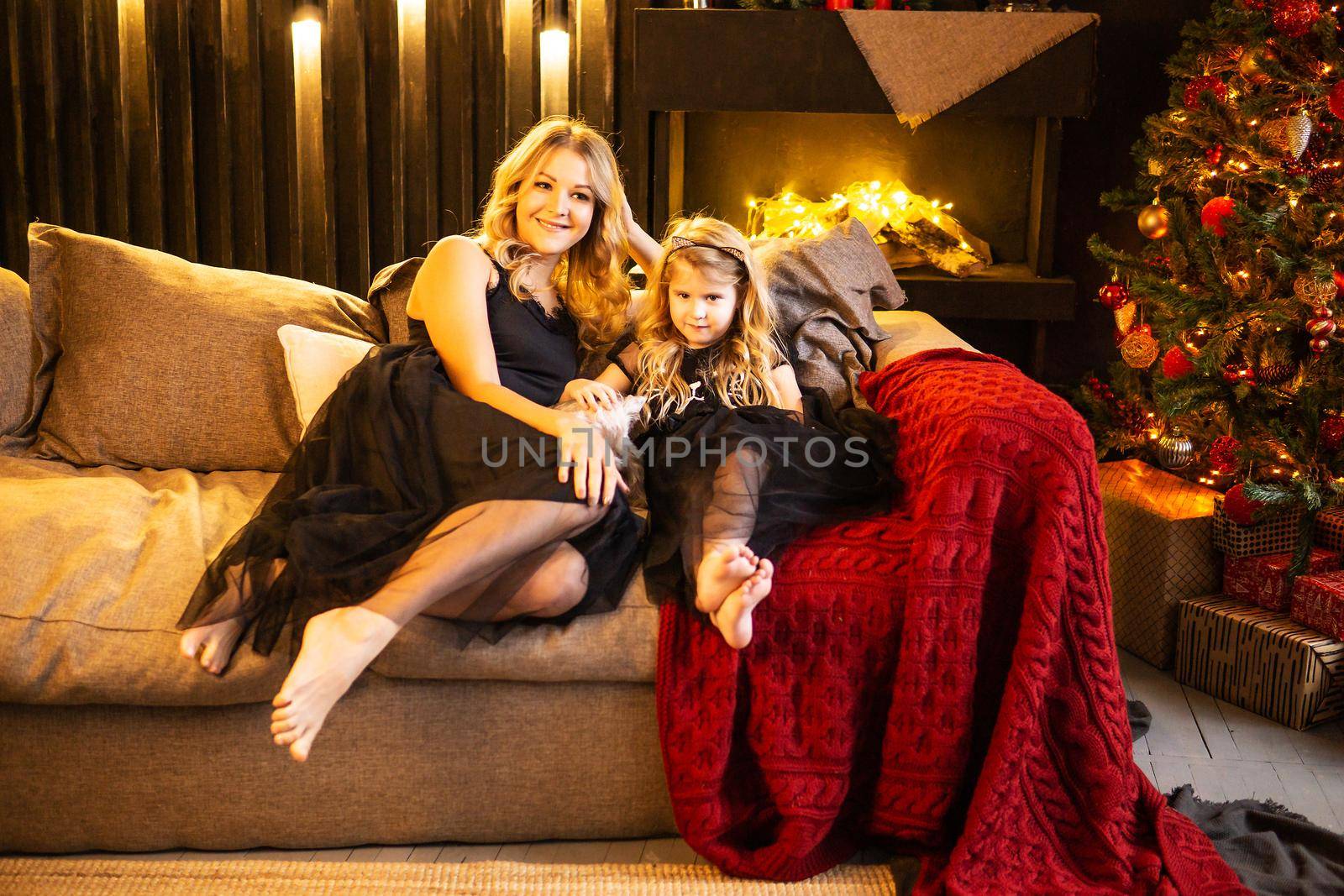 Mom and daughter in festive attire on the couch with their dog next to a Christmas tree decorated with garlands, balloons and Christmas toys. The concept of winter holidays is Christmas and New Year holidays. Magical festive atmosphere.