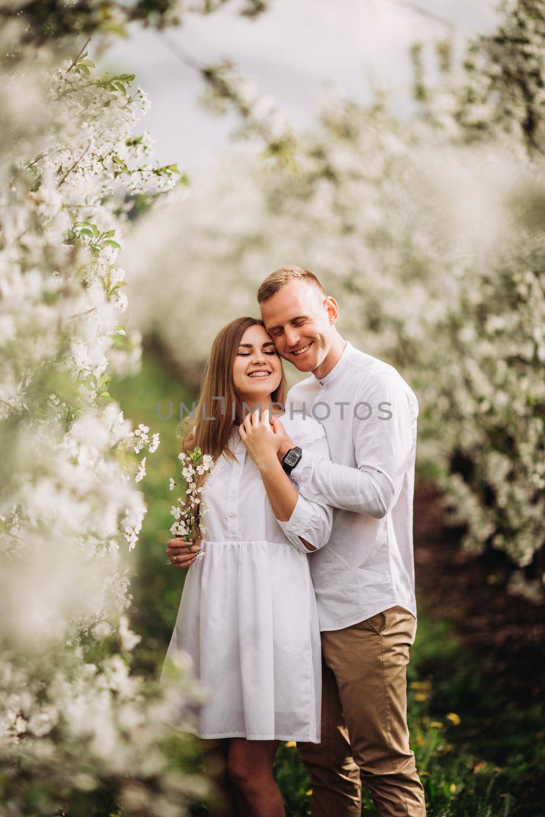 A happy young couple in love stands in a garden of blooming apple trees. A man in a white shirt and a girl in a white light dress are walking in a flowering park by Dmitrytph