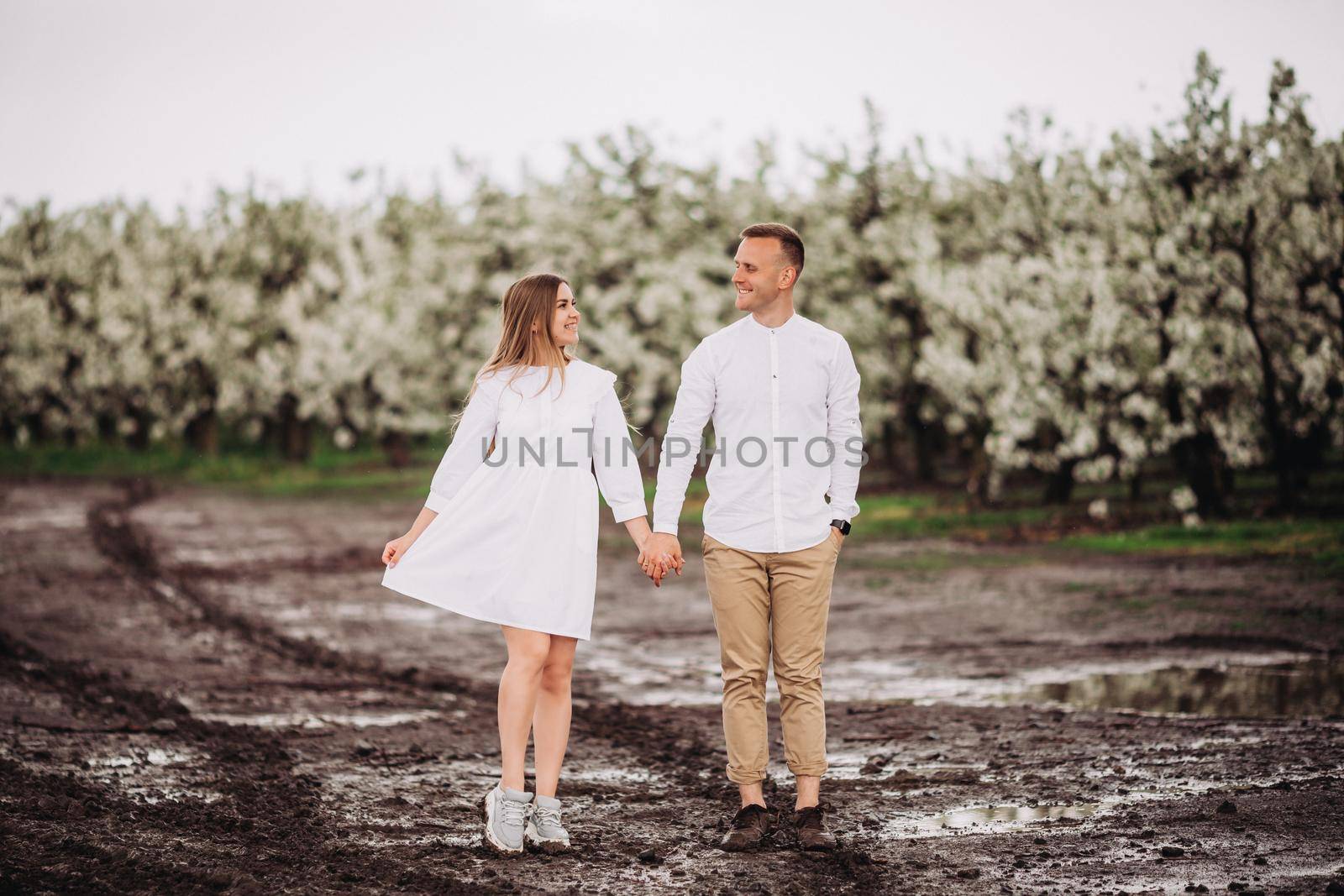 Happy family couple in the spring blooming apple orchard. Young couple in love enjoy each other while walking in the garden. Mud underfoot. The man holds the woman's hand. Family relationships by Dmitrytph