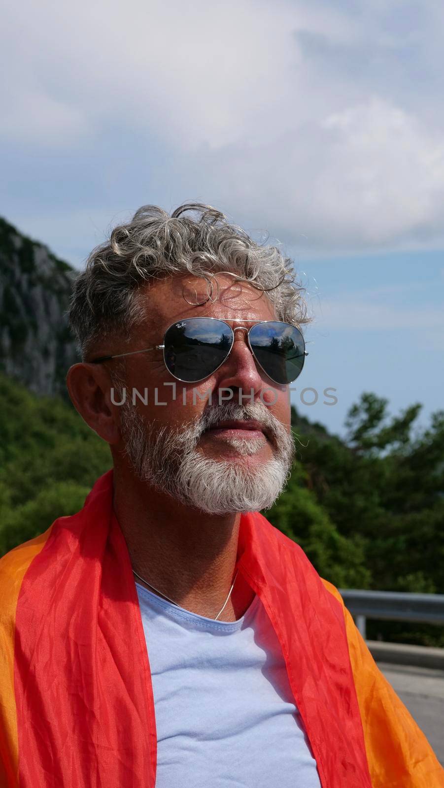 Portrait of a gray-haired senior elderly Caucasian man bisexuality with a beard and sunglasses with a rainbow LGBT flag in mountains. Celebrates Pride Month, Rainbow Flag Day