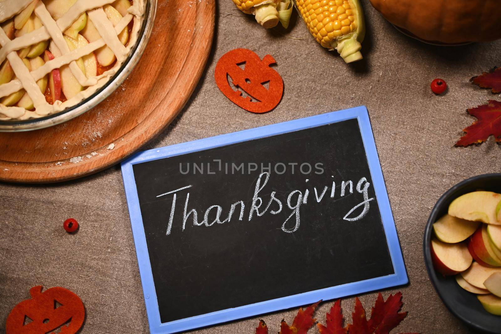 Flat lay of blackboard with chalk lettering Thanksgiving Day, next to a homemade festive pie and autumnal harvest of corn and pumpkin, on a table with linen tablecloth and dry fallen maple leaves