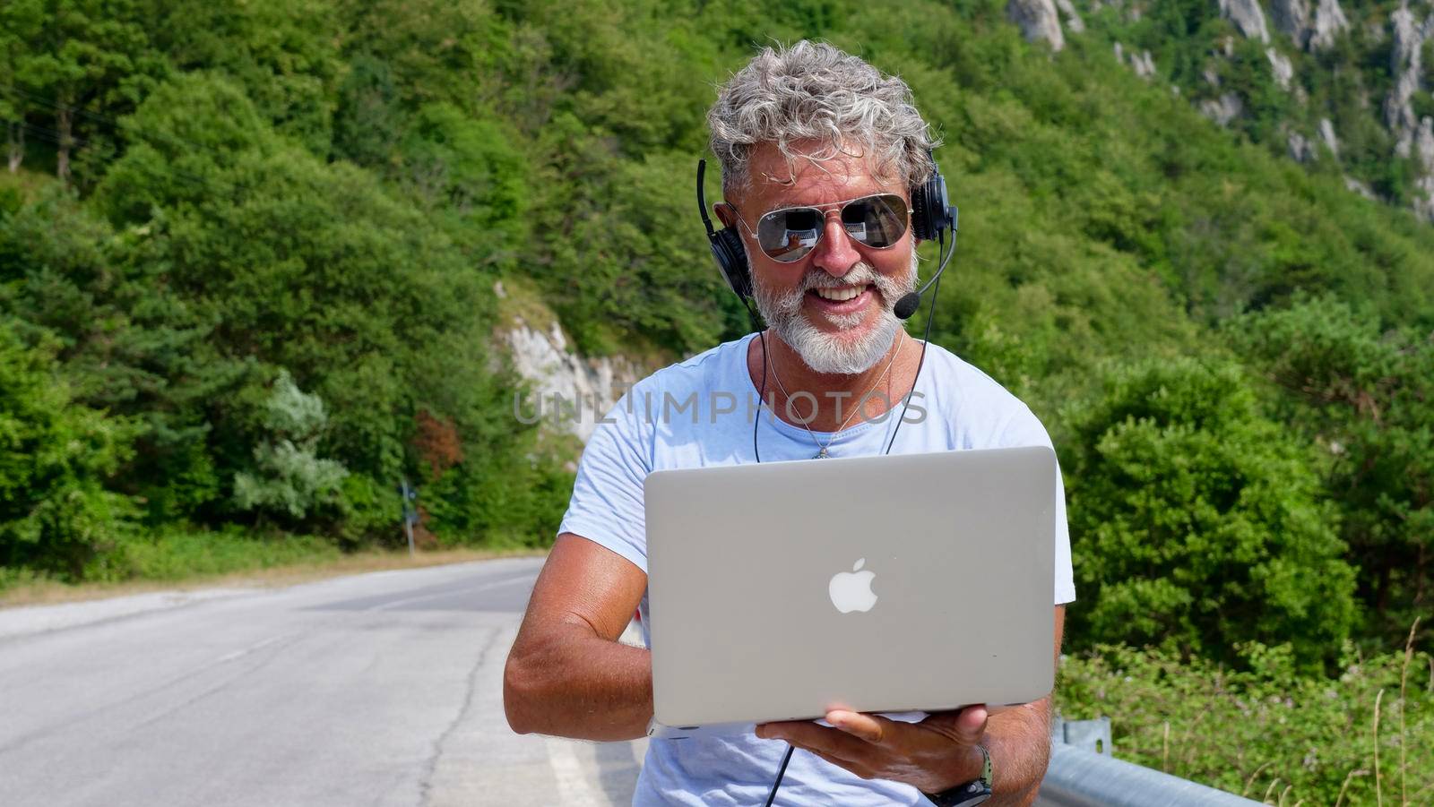 Gray-haired elderly man freelancer with a beard in sunglasses, working on a laptop on nature, road, mountains. A crazy and extraordinary old man sits on a yoga mat in the summer