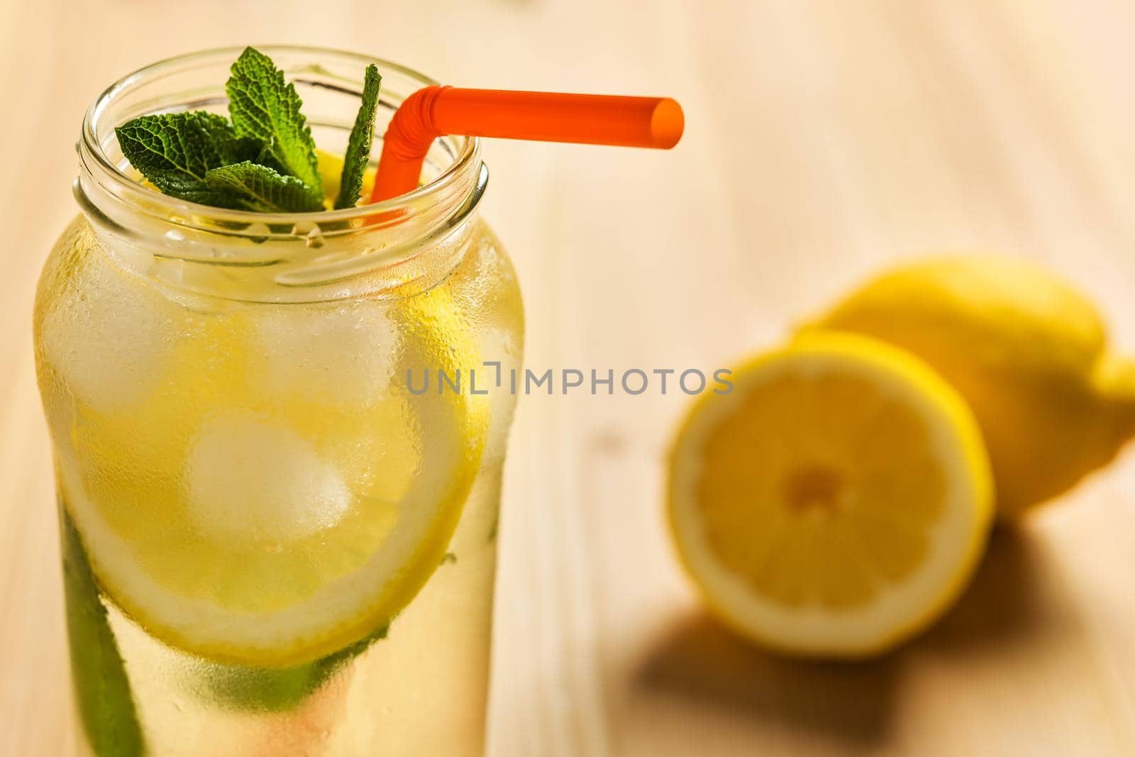 Close-up of the top of a glass jar with lemonade on a wooden table and some unfocused lemons in the background. The jug contains water, slices of lemon, ice, mint leaves and a cane to drink