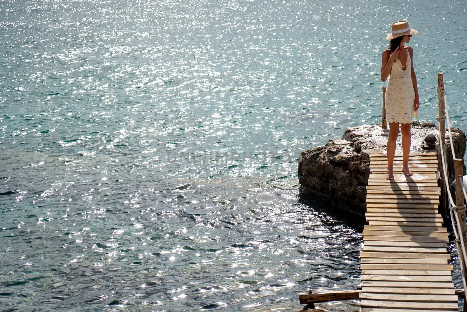 Brunette woman with hat and light beige dress walking on walkway over the sea