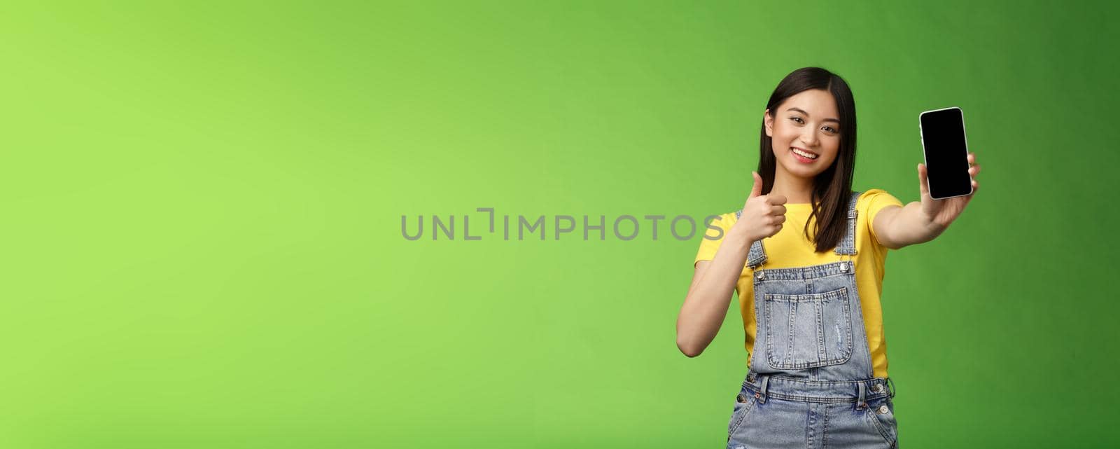 Cheerful cute asian woman judging good smartphone game, show thumb-up like sign, extend arm hold telephone screen, present awesome app, promoting application, stand green background.