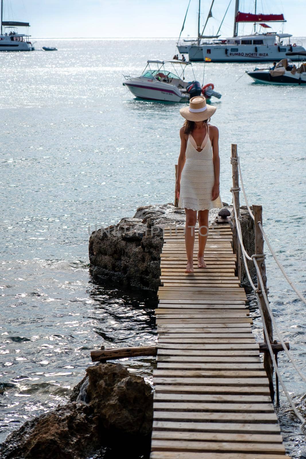 Brunette woman with hat and light beige dress walking on walkway over the sea