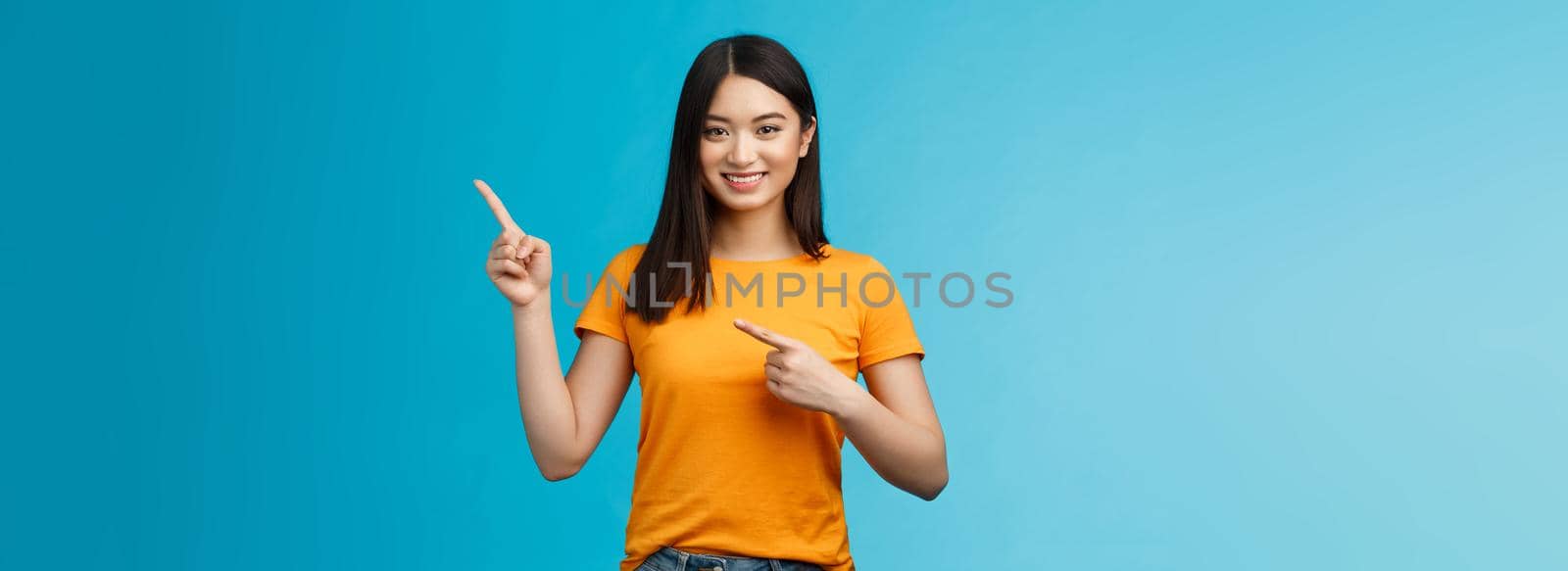 Friendly cheerful asian brunette girl inviting visit link interesting place, pointing upper right corner smiling joyfully camera, telling you about good promotion, stand blue background upbeat.