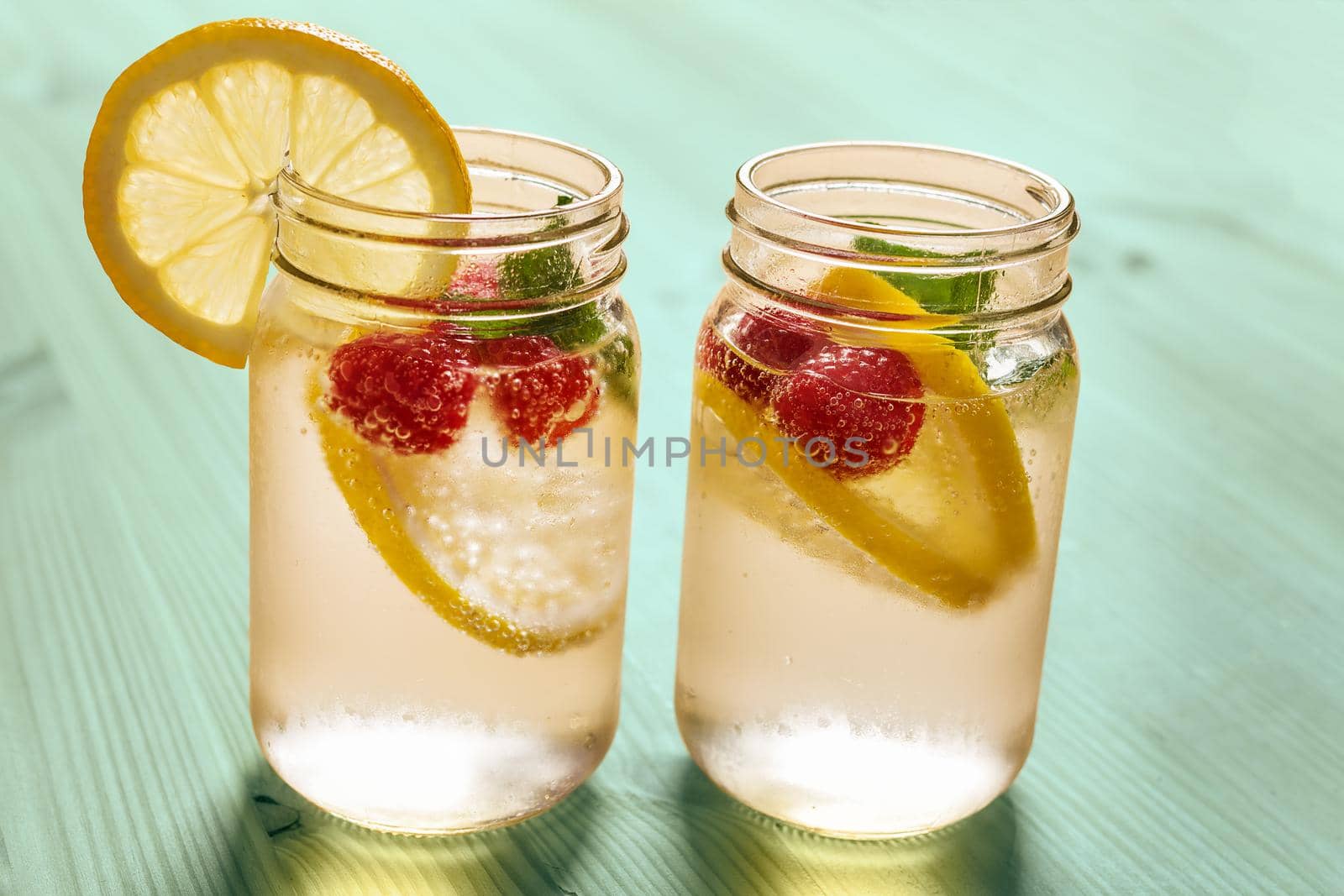 two glass jars with cold water, slices of lemon, mint and red berries, illuminated by sunlight on a green wooden table with some pieces of citrus, summer refreshments background, copy space