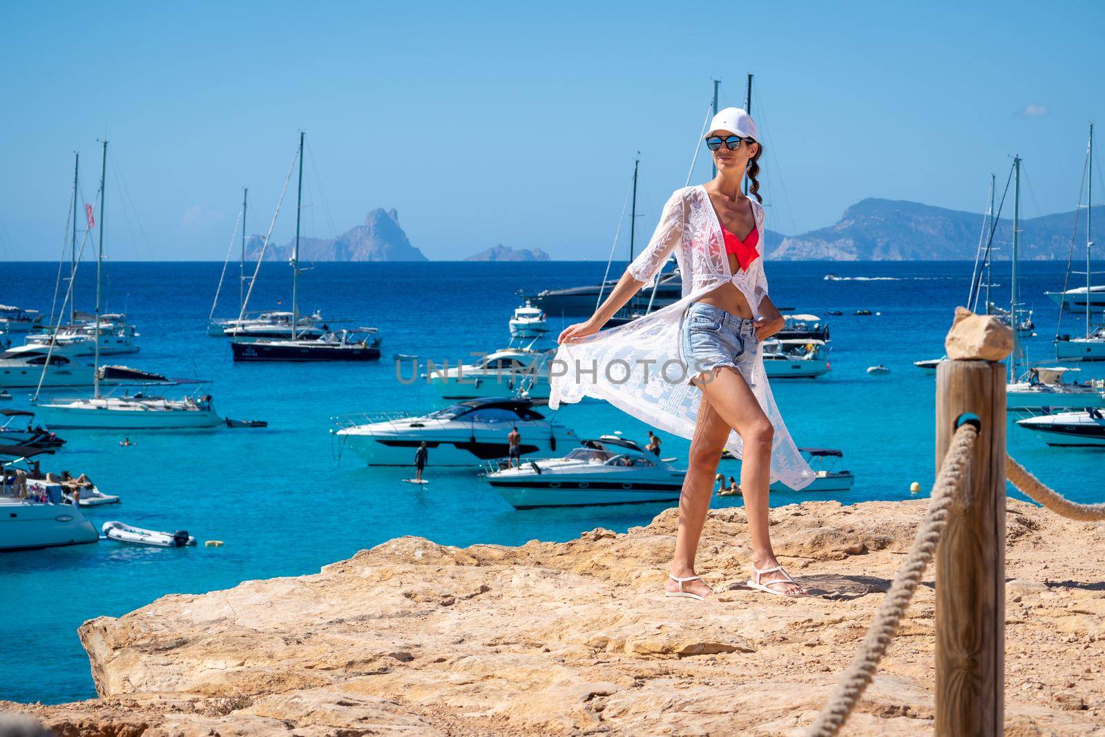 Brunette woman with white cap, crochet dress and shorts posing next to yachts in Formentera