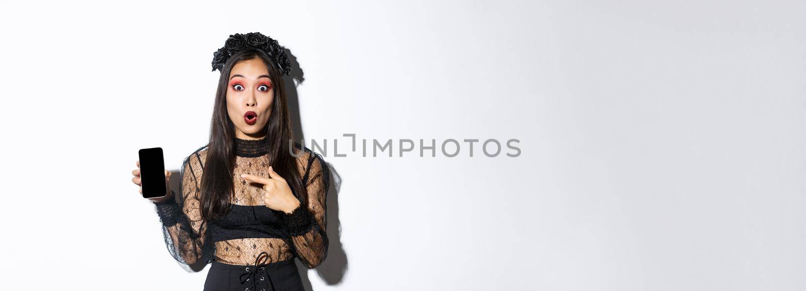 Surprised asian girl in black gothic dress with wreath, gasping amused and pointing finger at mobile phone display, showing halloween banner or promo, standing over white background.